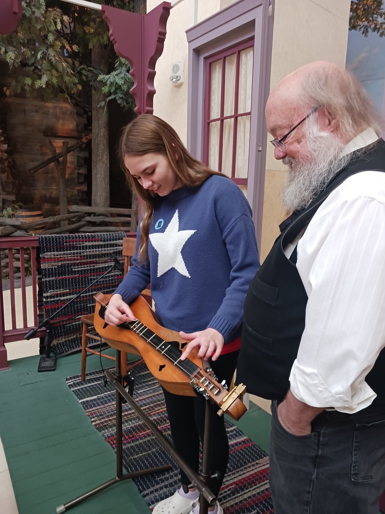An 8th grade student playing the dulcimer under the supervision of a museum guide.