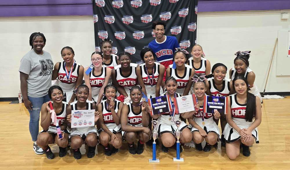 Group photo of the Hope High School cheerleading team posing indoors on a gym floor in front of a “National Cheerleaders Association” backdrop. The cheerleaders are wearing matching black, red, and white uniforms with “HOPE” and “CATS” lettering. They are holding trophies, medals, ribbons, and certificates. Two coaches or instructors stand on either side of the team, smiling.