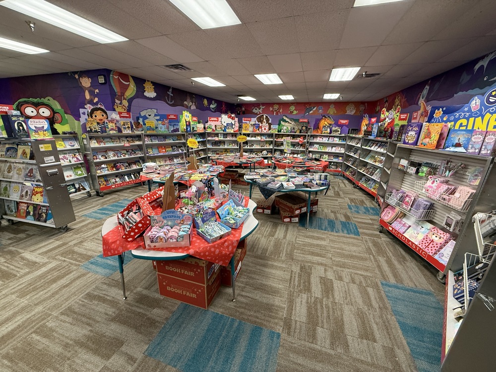 A school library with colorful book fair displays set up on tables. Bright signs, stacks of books, and decorations create a festive reading atmosphere.