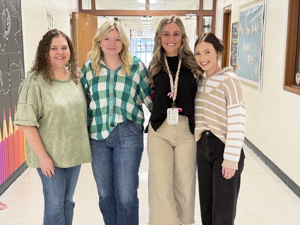 Four teacher residents stand together in a bright school hallway, smiling at the camera. They are dressed casually in jeans and sweaters, standing shoulder to shoulder. Behind them are colorful classroom bulletin boards and hallway decorations.