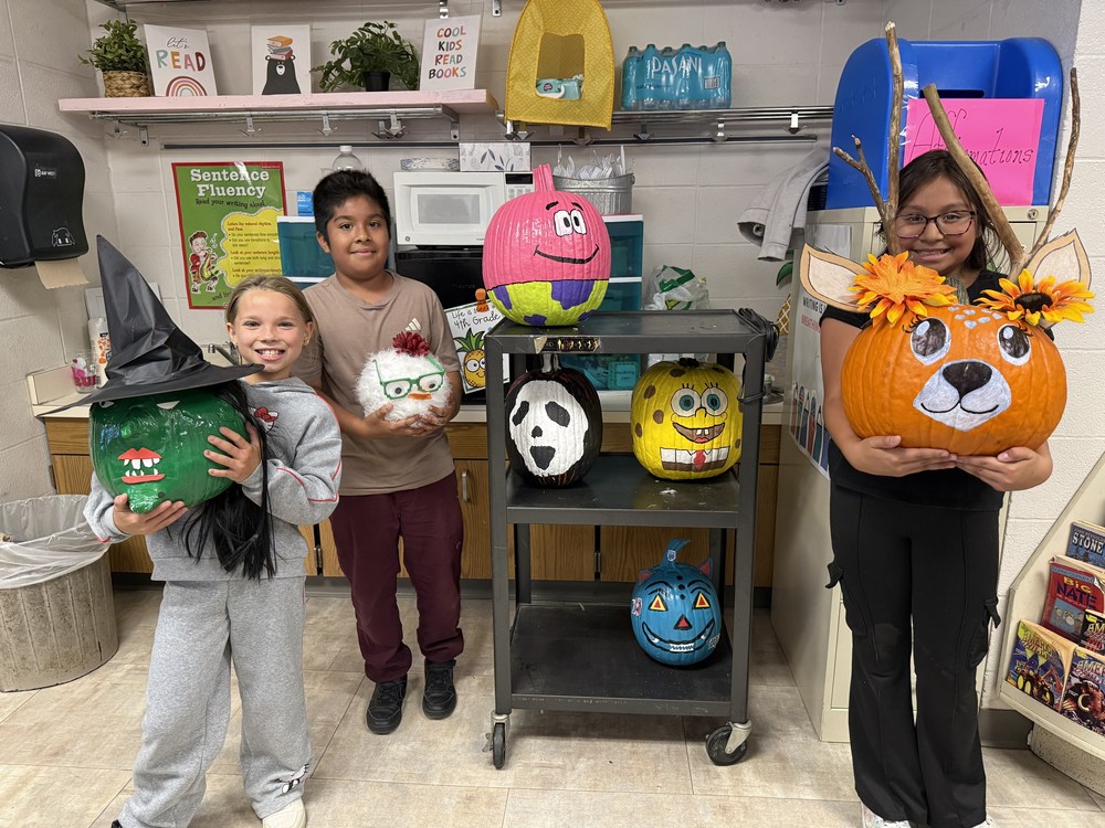 Three students stand in a classroom holding decorated pumpkins for a school pumpkin contest. One student on the left holds a green witch pumpkin with a hat and hair, the student in the middle holds a white fuzzy pumpkin with glasses, and the student on the right holds an orange pumpkin decorated like a deer with antlers and sunflowers. A cart behind them displays more painted pumpkins, including a pink pumpkin with a smiling face, a panda, a black and white pumpkin, a SpongeBob design, and a blue pumpkin with a jack-o’-lantern face. The classroom has posters, books, and plants in the background.