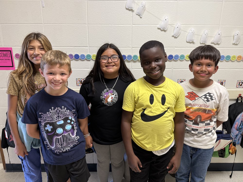 Five elementary students stand side by side in a school hallway, smiling at the camera. Behind them is a classroom wall decorated with colorful smiley face cutouts and white ghost decorations. Each student wears a different graphic T-shirt, including designs with a smiley face, video game controller, and cars. They appear cheerful and proud, representing leadership in their class.
