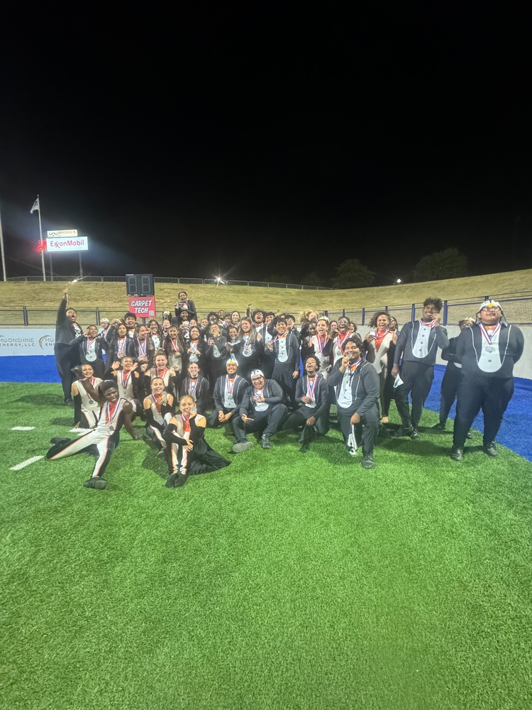 A large group of smiling high school band members and color guard students from Hope High School pose together on a football field at night after a performance. They are wearing matching uniforms and medals around their necks, celebrating their achievement under the stadium lights. The group stands and kneels on bright green turf with a dark sky above and a few lit sponsor signs visible in the background.