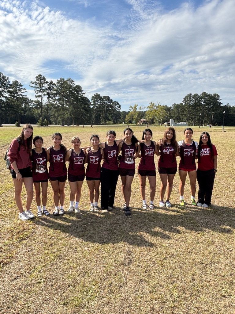 A group of ten female cross country runners and their coach from Hope High School stand in a line on a grassy field, smiling at the camera. They are wearing matching maroon uniforms with the school’s logo and sandals or running shoes. Tall pine trees and a blue sky with scattered clouds are visible in the background.