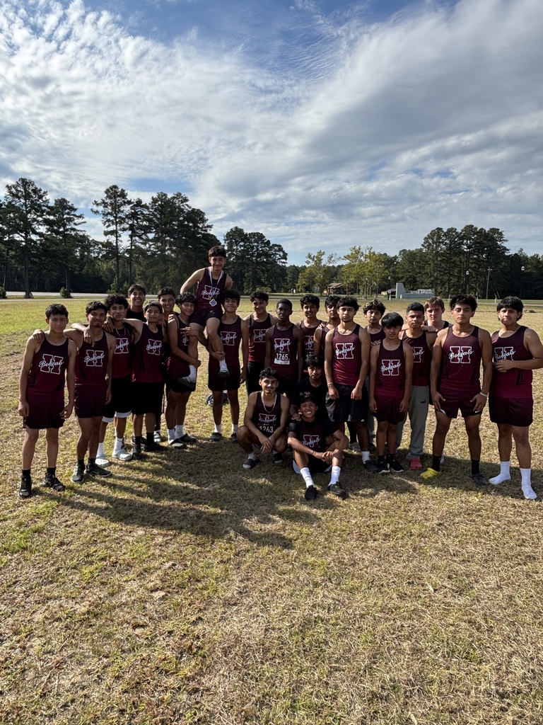 A large group of male cross country runners from Hope High School gather together on a grassy field for a team photo. Many are standing shoulder to shoulder, while a few are kneeling or sitting in front. They are wearing matching maroon uniforms with the school’s logo. The background shows trees, a clear blue sky, and scattered clouds.