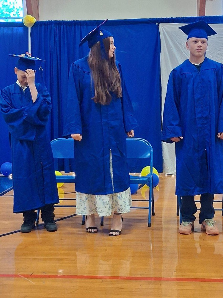 Three students in graduation uniforms standing in front of their chairs