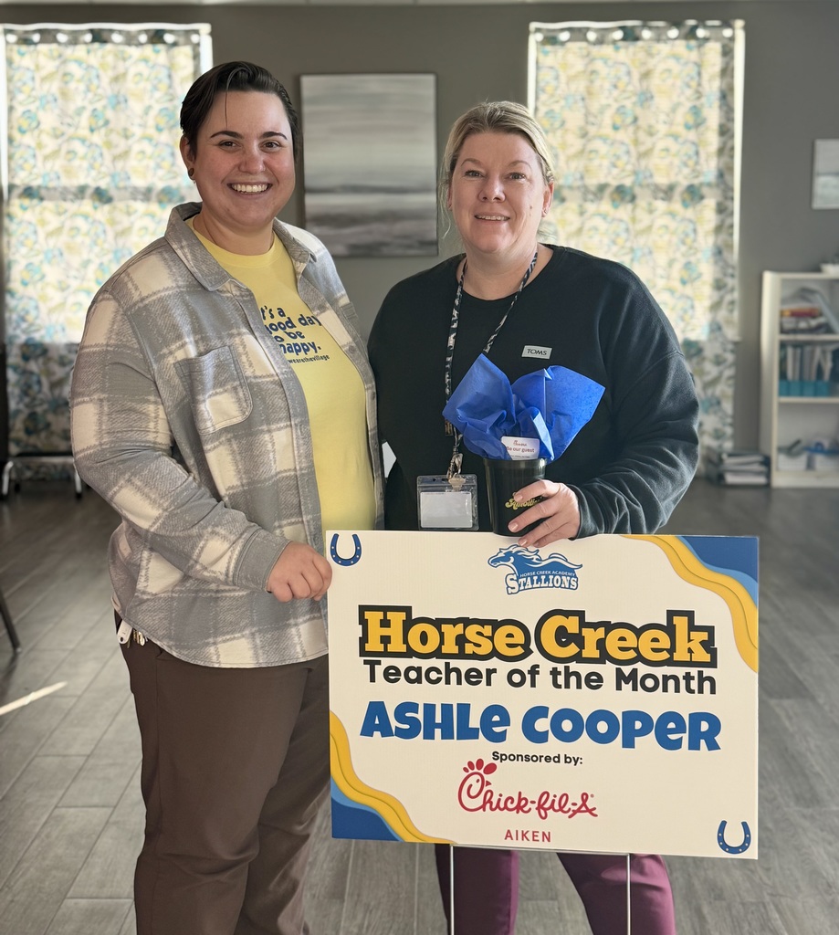 Two women holding a sign for Horse Creek Teacher of the Month, with one wearing a yellow shirt and the other in a black shirt.