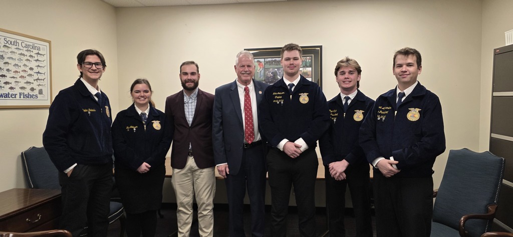 A group of people in jackets and ties, standing in an office. One person wears a red tie.