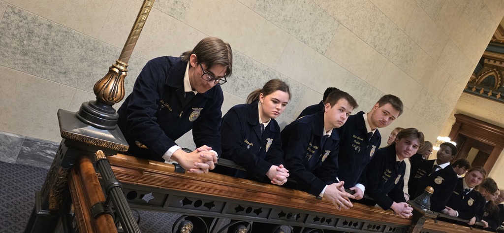 A group of students in uniforms leaning on a balcony. The background shows a room with a decorative railing.
