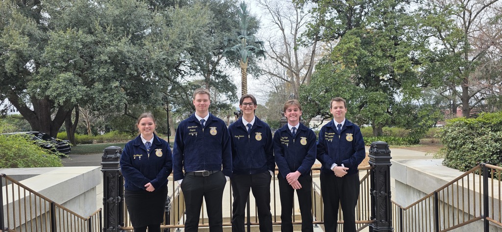 Five individuals in matching uniforms pose near a metal railing. Behind them, greenery and trees are visible.