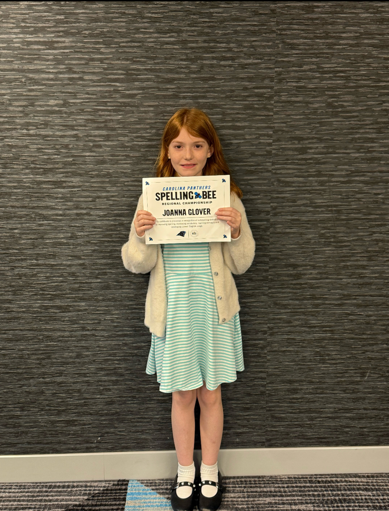 Girl in blue dress holds certificate with name and achievement, standing in front of a gray textured wall.
