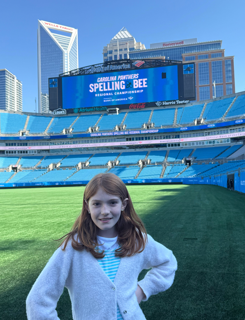 Girl stands in empty stadium with large screen displaying "Carolina Panthers Spelling Bee" and "Regional Championship."