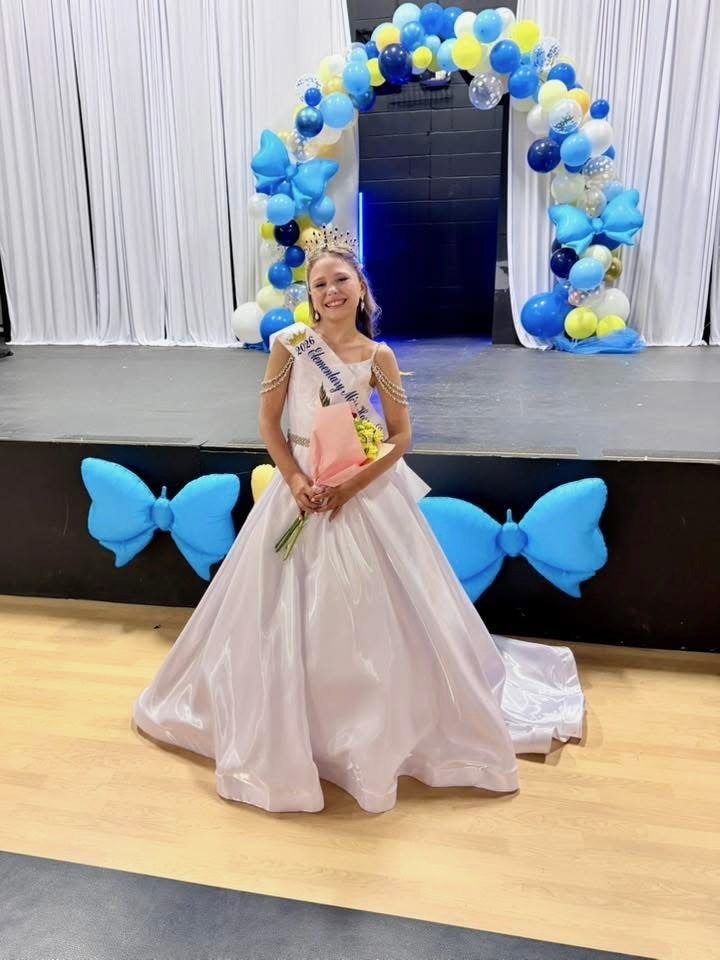 A woman in a white gown stands on stage with a crown, holding a bouquet, surrounded by colorful balloons.