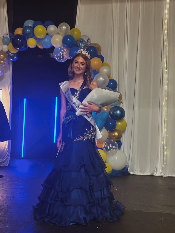 A woman in a blue dress stands under a balloon arch, holding a bouquet, with a sash around her waist.