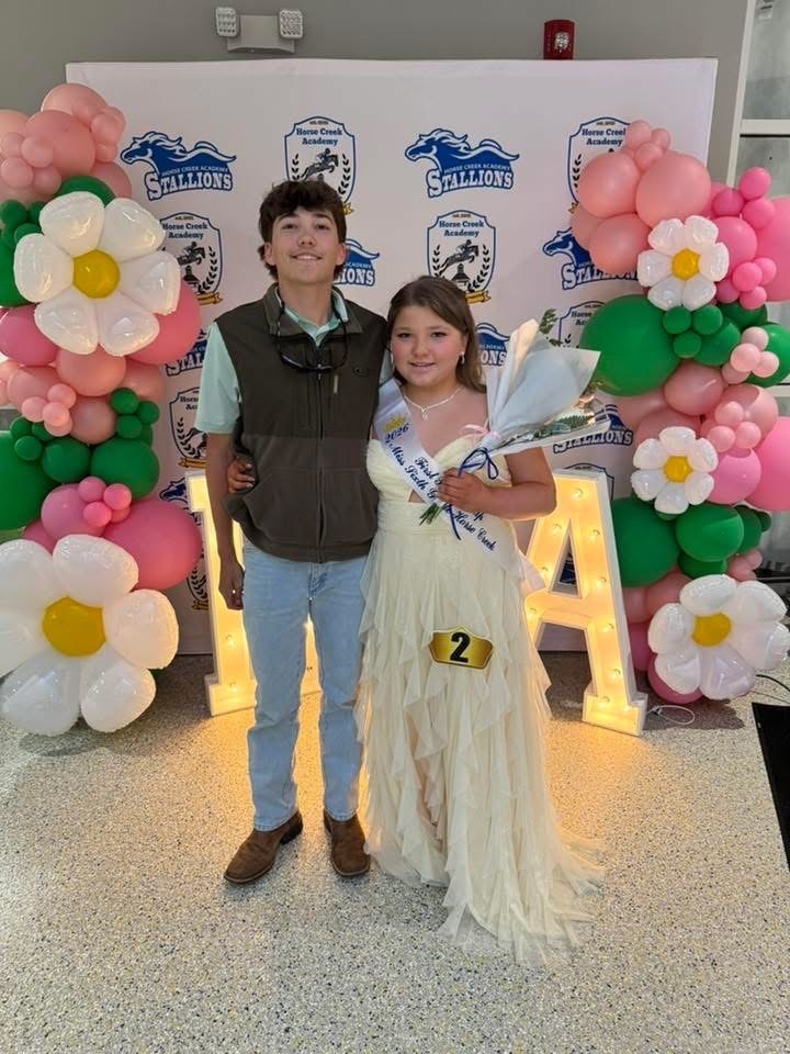 A boy and girl stand next to a floral arch and letters. The boy wears a vest, and the girl holds flowers.