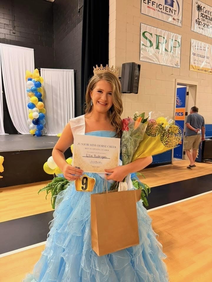 A woman in a blue dress and crown holds a certificate and bouquet, standing in a gymnasium.
