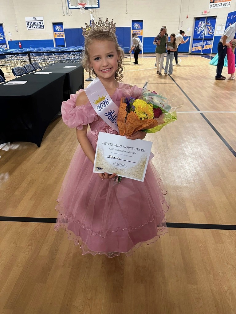 A girl in a pink dress holds a bouquet and a certificate, smiling in a gym. Other people walk around in the background.
