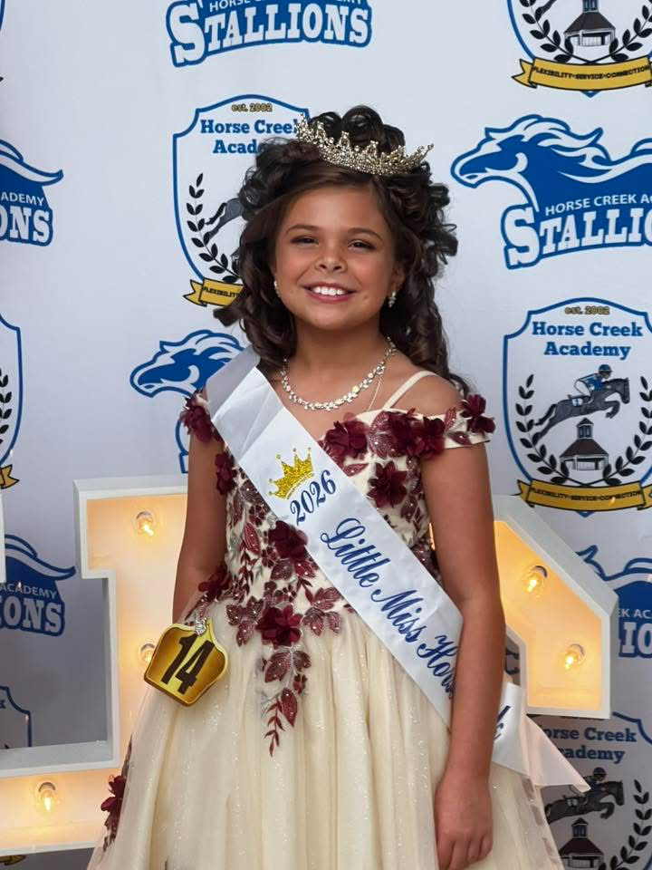 A girl in a yellow gown, a crown, and a sash saying "Little Miss Horse" stands in front of a white backdrop.