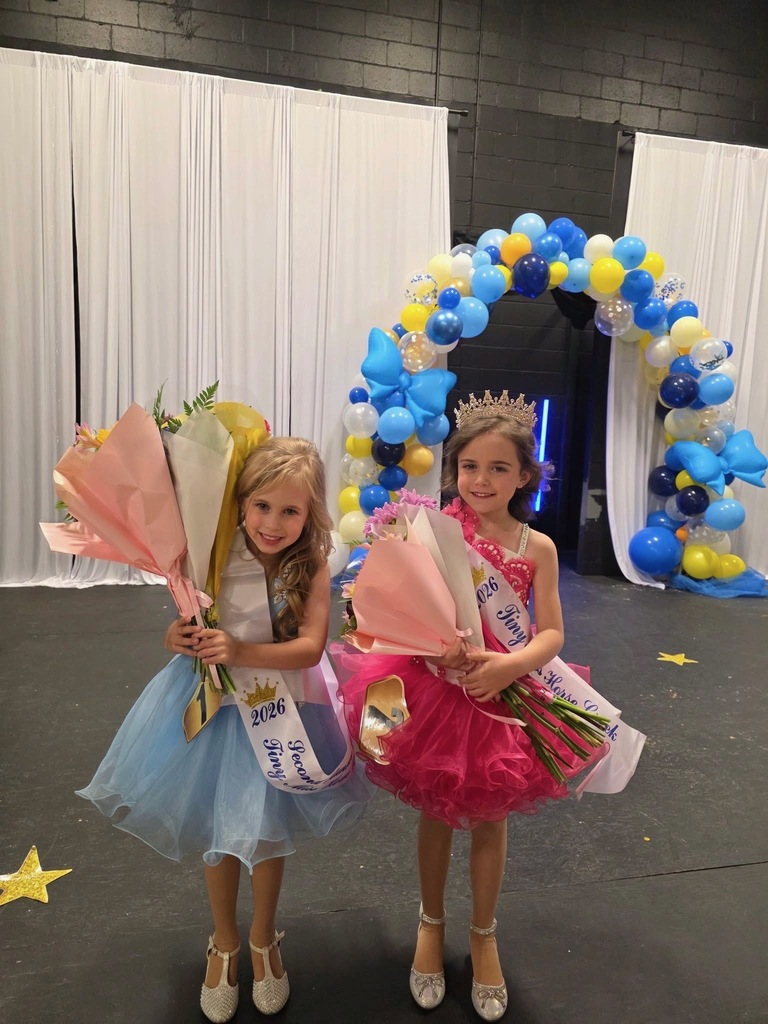 Two young girls in colorful dresses and crowns hold flower bouquets in front of a balloon arch.