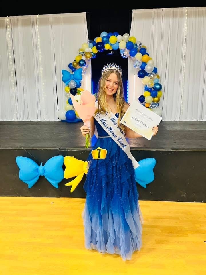Woman in a blue gown and sash holding a bouquet and certificate on a stage with balloons.