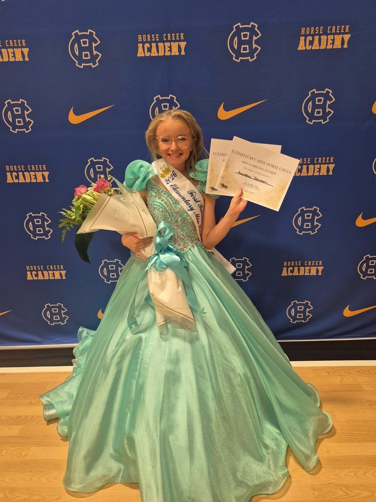A girl in a blue gown stands holding certificates and flowers. Behind her is a blue backdrop with "Horse Creek Academy" in white text.