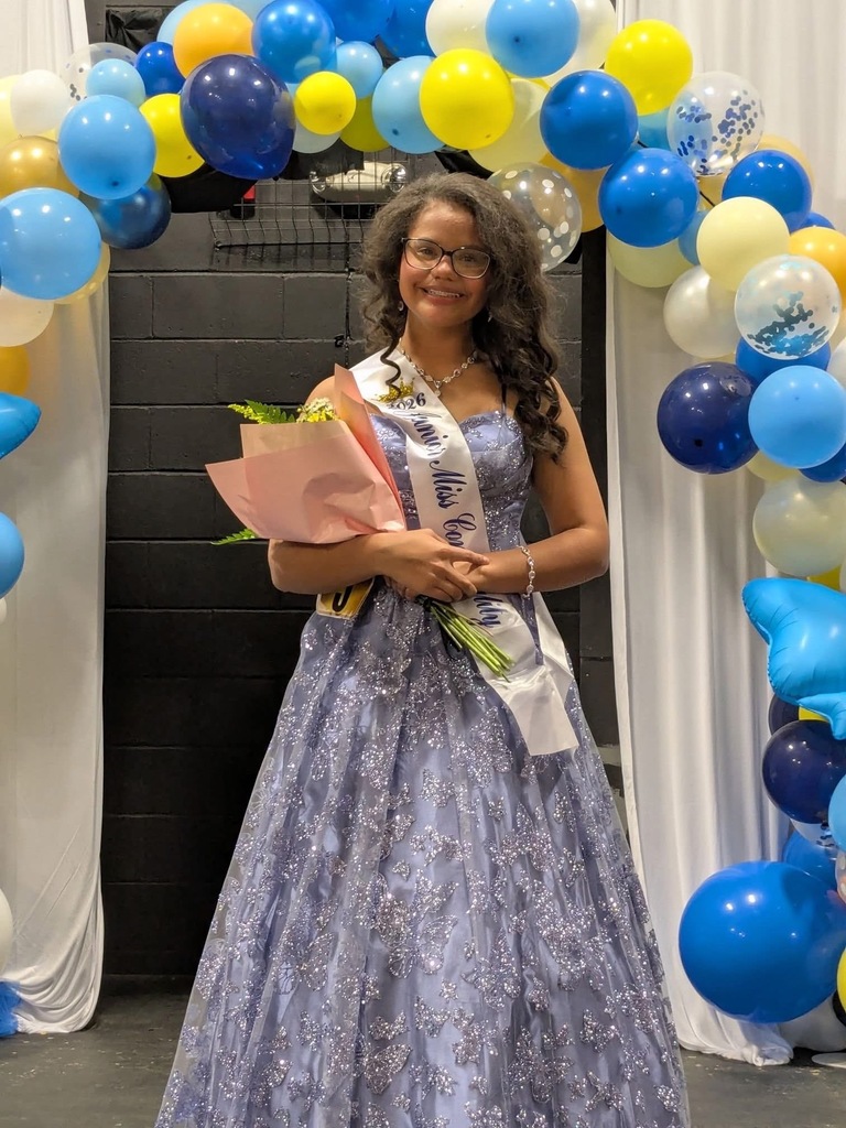 A woman in a blue dress, holding flowers and a sash, stands in front of a colorful balloon arch.