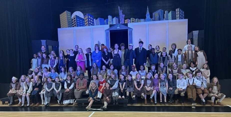 A group of people in costumes pose for a photo on a stage with a city backdrop.