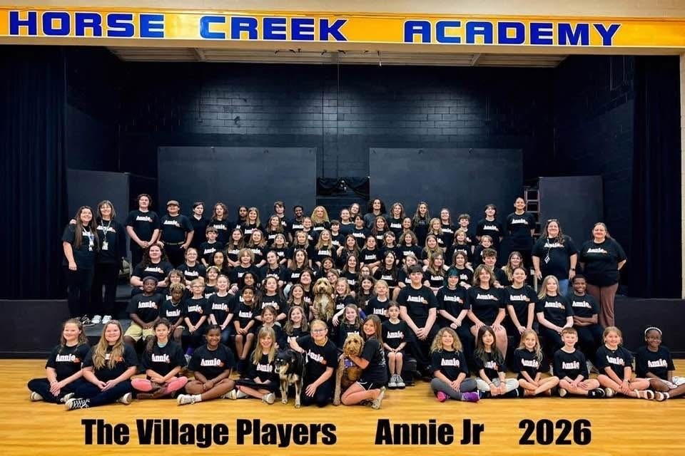A group of people wearing black t-shirts, sitting and standing, on a stage, with "The Village Players Annie Jr 2026" text.