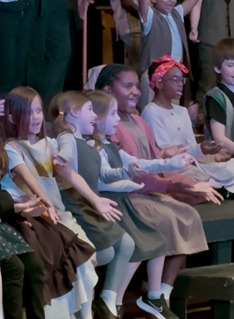 Children in vintage clothing, seated and standing, with two adults behind them. Some children wear glasses.