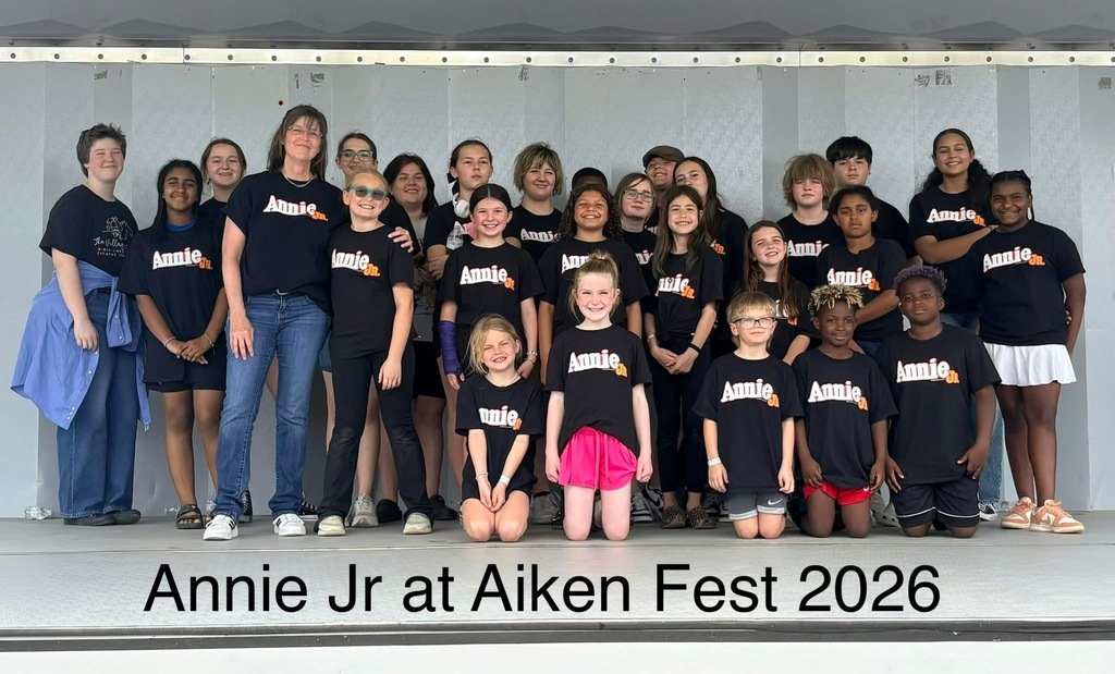 Group photo at an event: children and adults in black shirts, some wearing glasses, with text "Annie Jr at Aiken Fest 2026".