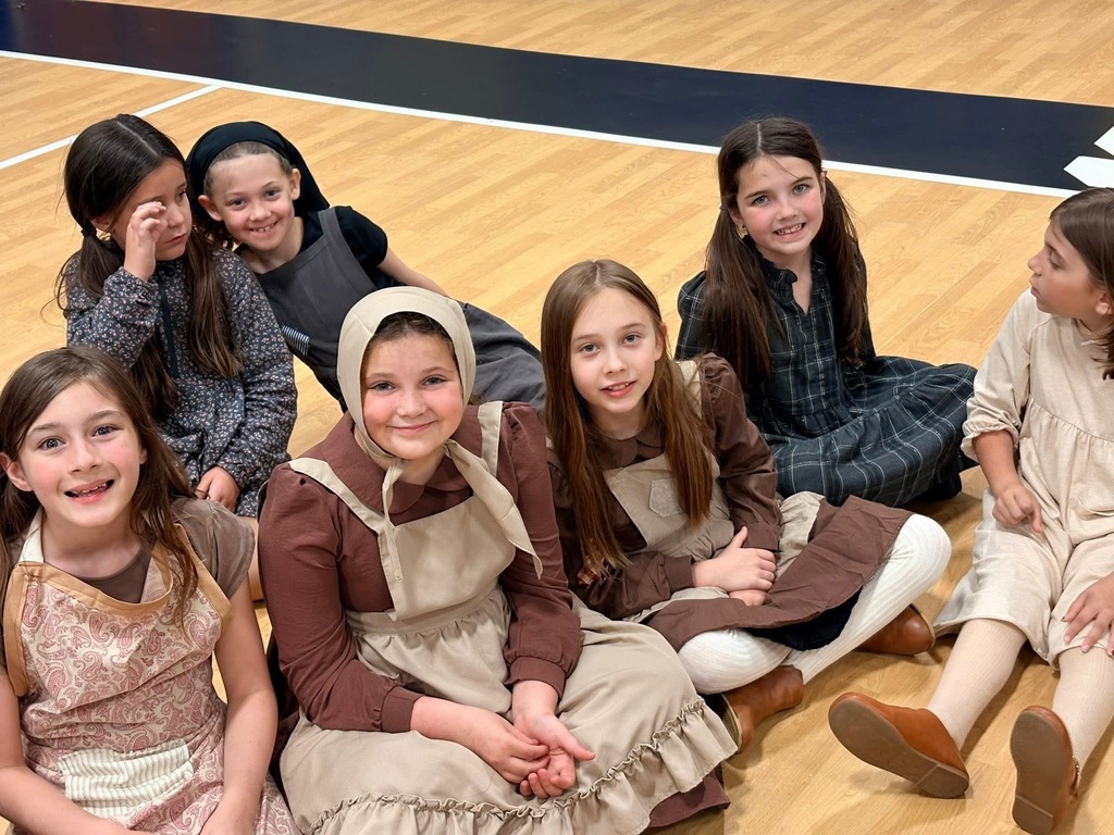 A group of young girls dressed in historical clothing, sitting in a circle on a gym floor.