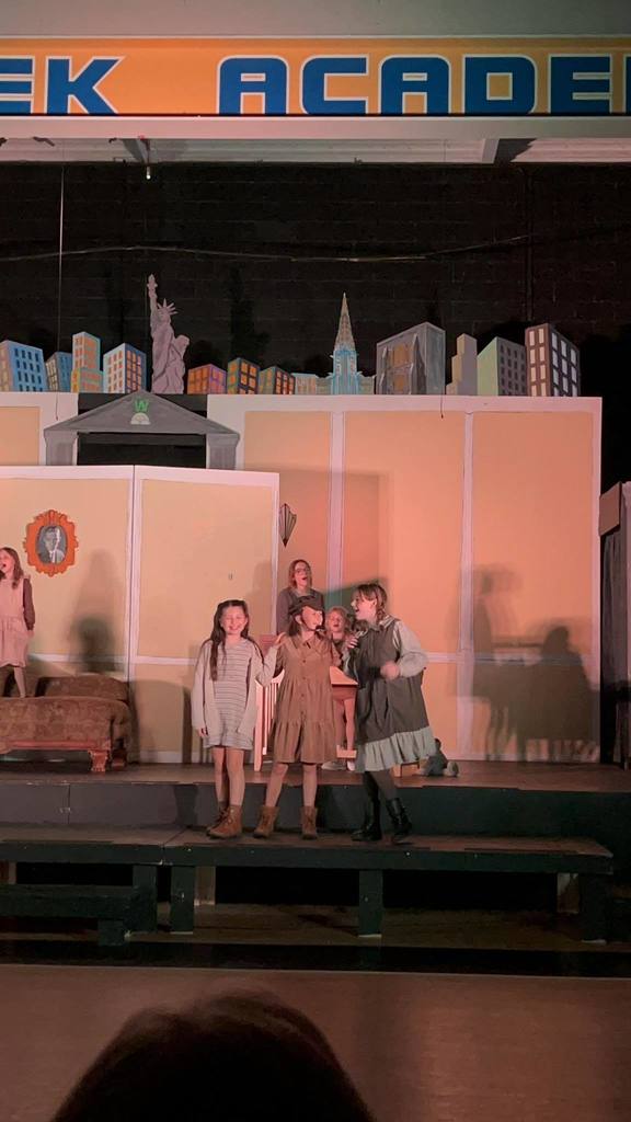 A group of children perform a play on a stage with a backdrop of city buildings.