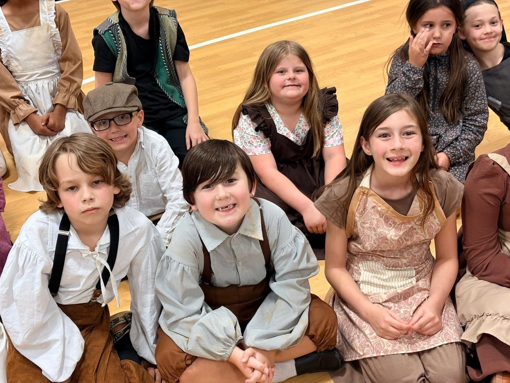 Several young students, wearing historical costumes, pose for a photo on a gymnasium floor. Some have hats, and they sit on the floor with smiles.