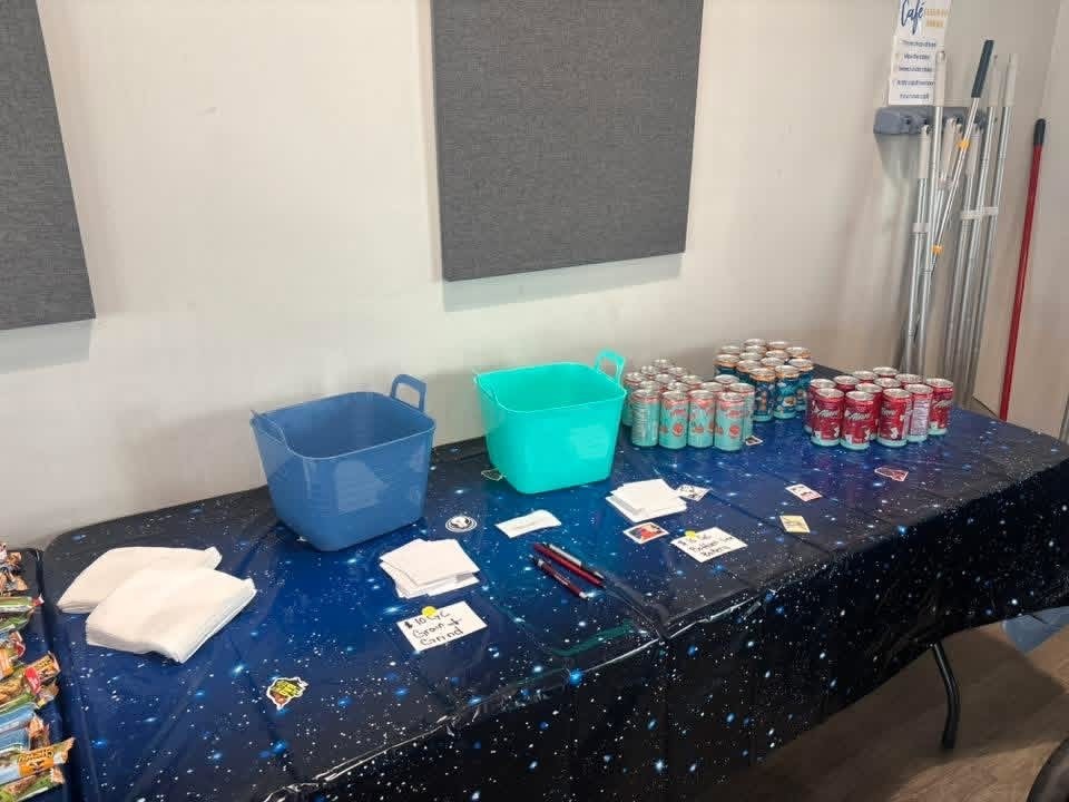 A table with a blue tablecloth, a blue bucket, a green bucket, cans, cards, and napkins.