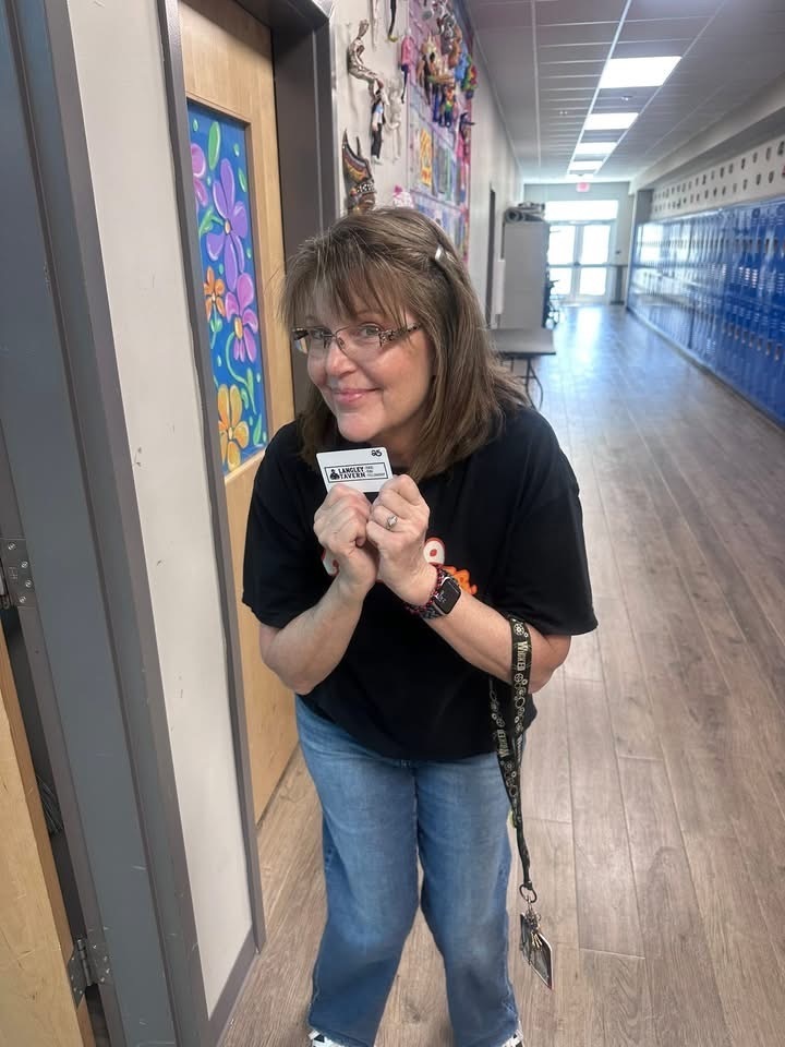 A woman in a black shirt holds a small camera while standing in a school hallway with lockers.