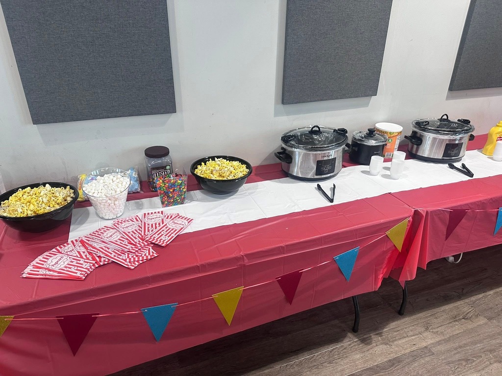 A long table covered with a red tablecloth and colorful triangular decorations. Bowls of popcorn, candy, and a crockpot.
