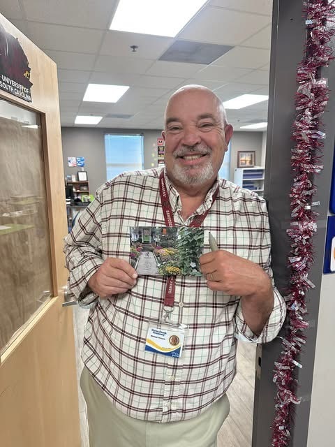 A man in a plaid shirt and ID badge stands next to a door holding a card. Behind him is a room with shelves, a window, and decorative red and white streamers.
