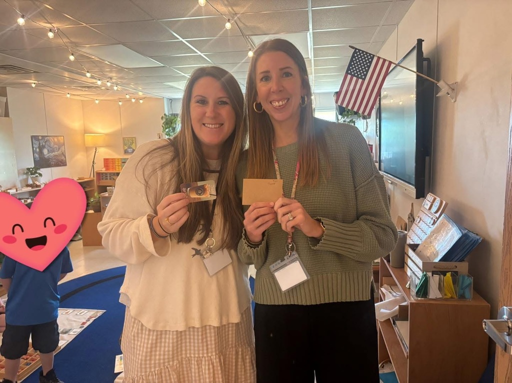Two women stand, one holding a card, in a classroom with a flag, books, and a smiling boy.