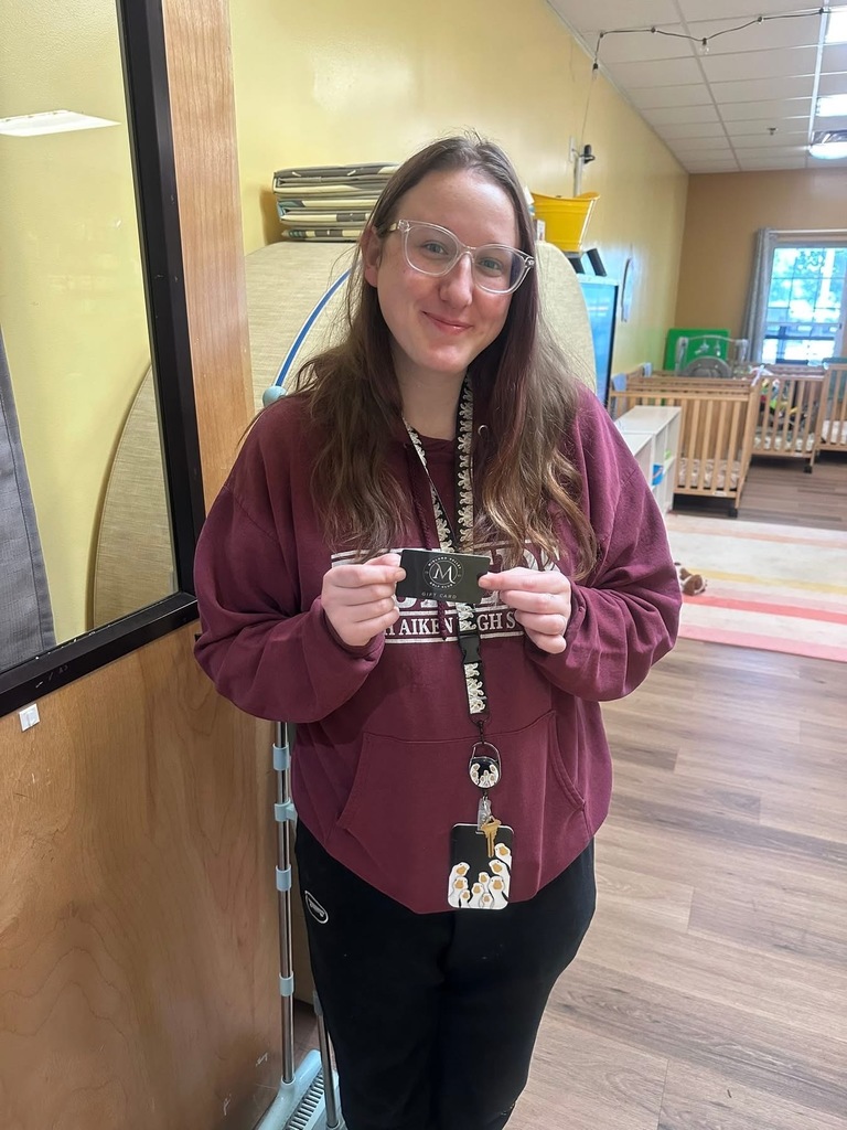 A woman in a maroon hoodie holds a badge and smiles at the camera. Behind her is a room with wood floors and a crib.