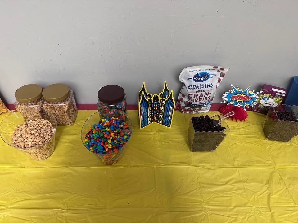 A table covered with a yellow cloth, holding various snacks and containers, including popcorn, candies, and a bag of raisins.