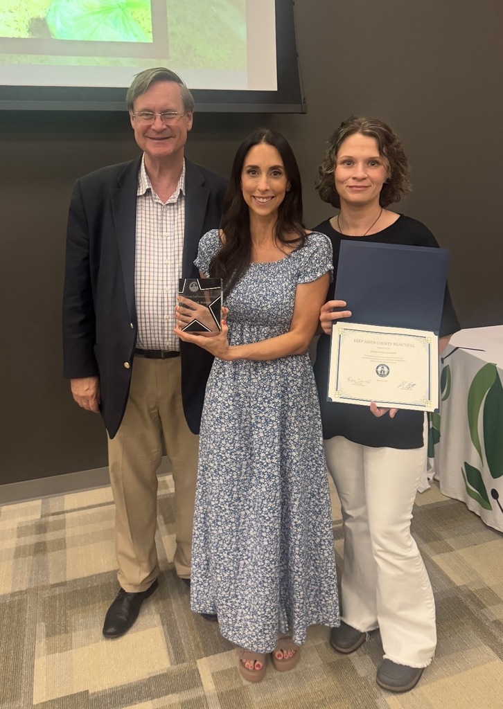 Three people stand together, smiling. A woman holds a certificate and an award. The man beside her wears glasses.