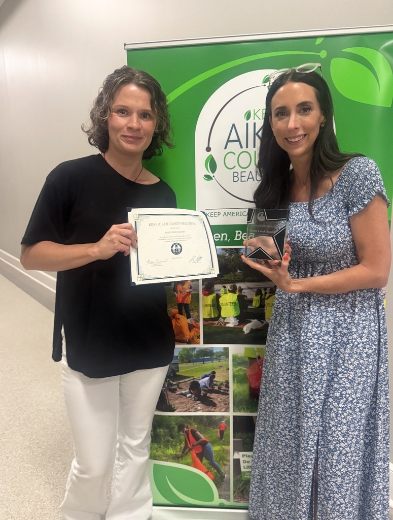 Two women hold awards in front of a banner with green text and photos of people.