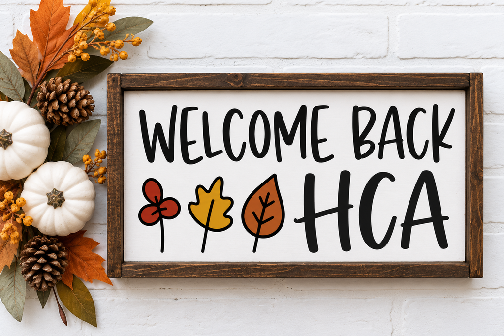 A white brick wall with a wooden sign reading "Welcome Back HCA" in black. Leaves and small pumpkins are beside the sign.