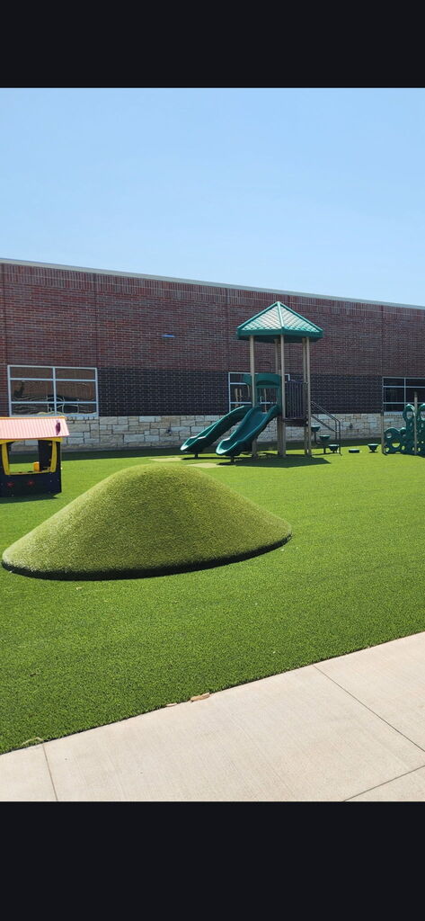 Three photos show the new Pre-K and Kindergarten playground at Summit Hill Elementary. The playground features bright green artificial turf, multiple play structures with slides and climbing features, a colorful playhouse, dome climbers, a grassy play mound, and a spinning seat. The playground is surrounded by the brick exterior of the school’s new construction under a clear blue sky.