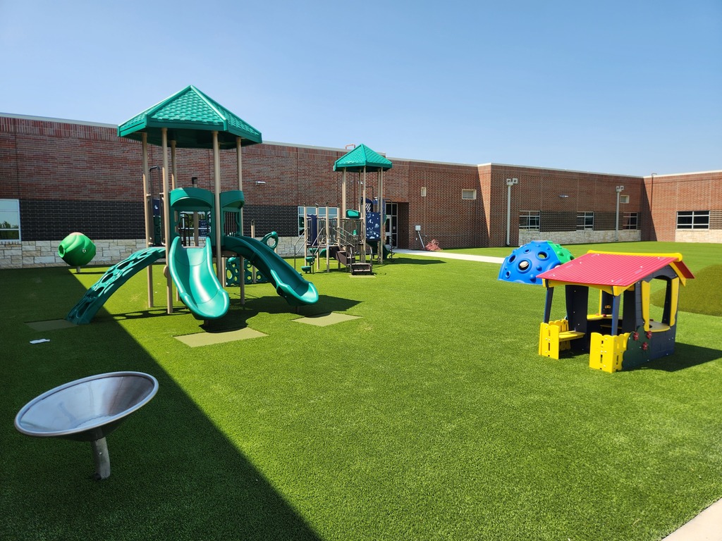 Three photos show the new Pre-K and Kindergarten playground at Summit Hill Elementary. The playground features bright green artificial turf, multiple play structures with slides and climbing features, a colorful playhouse, dome climbers, a grassy play mound, and a spinning seat. The playground is surrounded by the brick exterior of the school’s new construction under a clear blue sky.