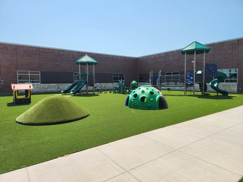 Three photos show the new Pre-K and Kindergarten playground at Summit Hill Elementary. The playground features bright green artificial turf, multiple play structures with slides and climbing features, a colorful playhouse, dome climbers, a grassy play mound, and a spinning seat. The playground is surrounded by the brick exterior of the school’s new construction under a clear blue sky.