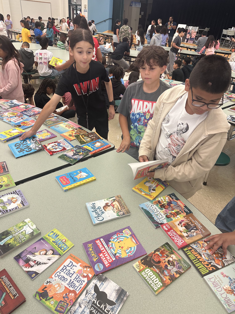 Three students stand at a cafeteria table filled with a wide variety of chapter books and graphic novels.