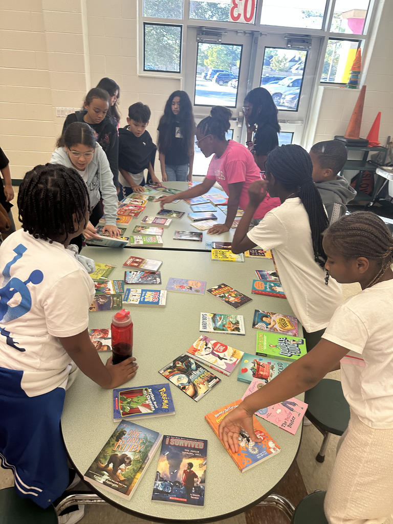 A group of students gathers around several cafeteria tables covered with books.