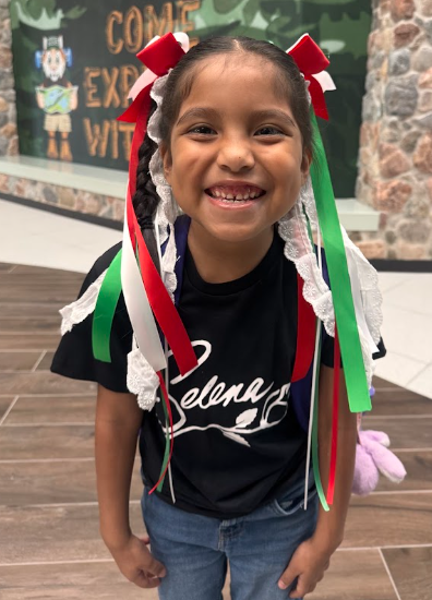 A student in a black SelenaT-shirt and jeans smiles. She wears a red and white ribbon headband with green and white ribbons for Boots and Ribbons Day.