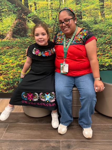 Ms. Nambo and her daughter sit together on a chair in front of a forest mural.